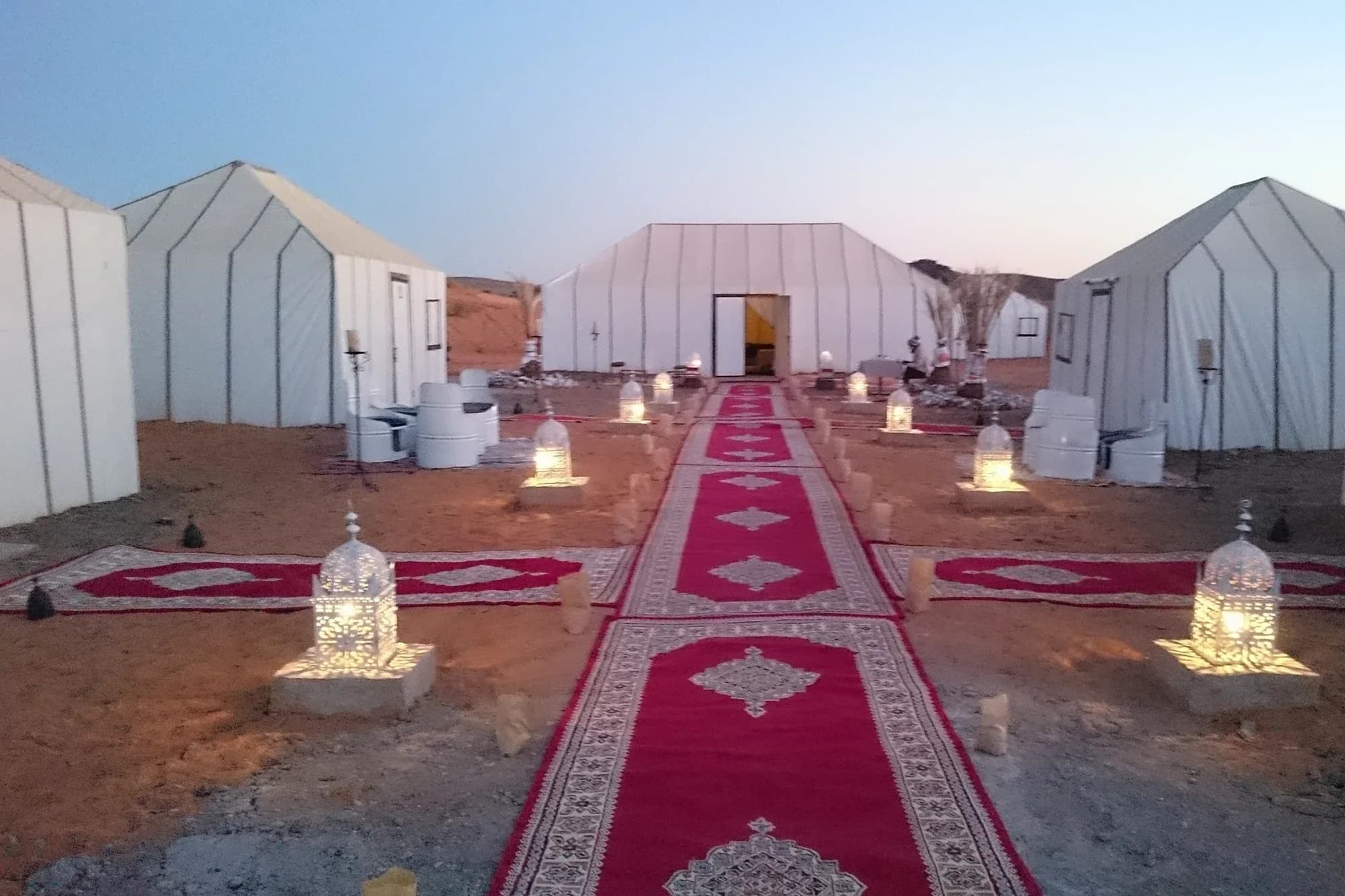 Campamento de jaimas blancas en el desierto marroquí con alfombras y farolillos al atardecer